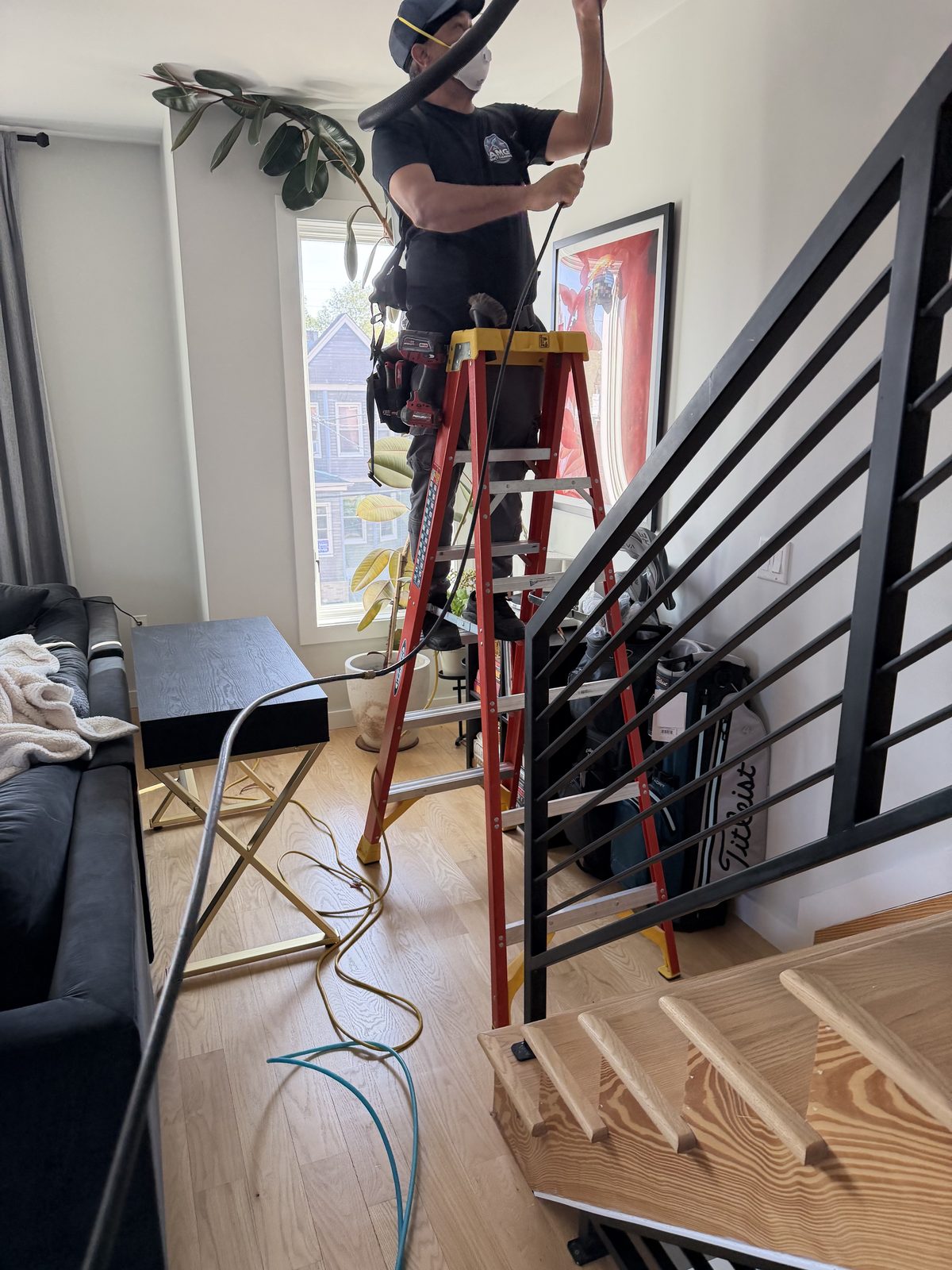 An AMG technician on a step ladder at a ceiling vent, the vacuum hose run up beside him