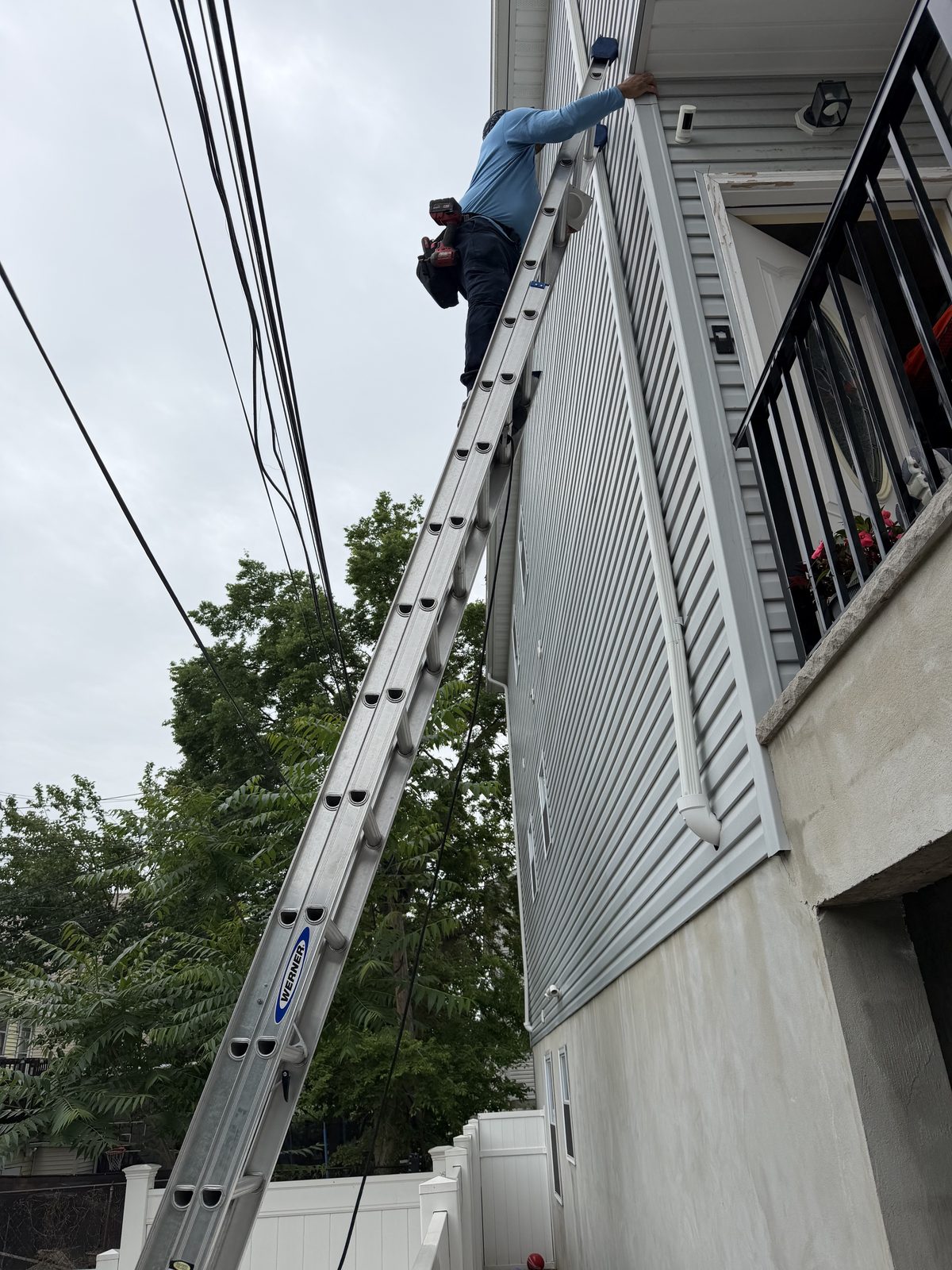 An AMG technician at the top of an extension ladder, working at a vent beside a balcony
